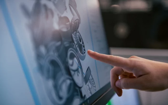 A imagem possui uma tela de computador com algumas peças que serão impressas em 3D; a mão de uma pessoa está apontando para esta tela.