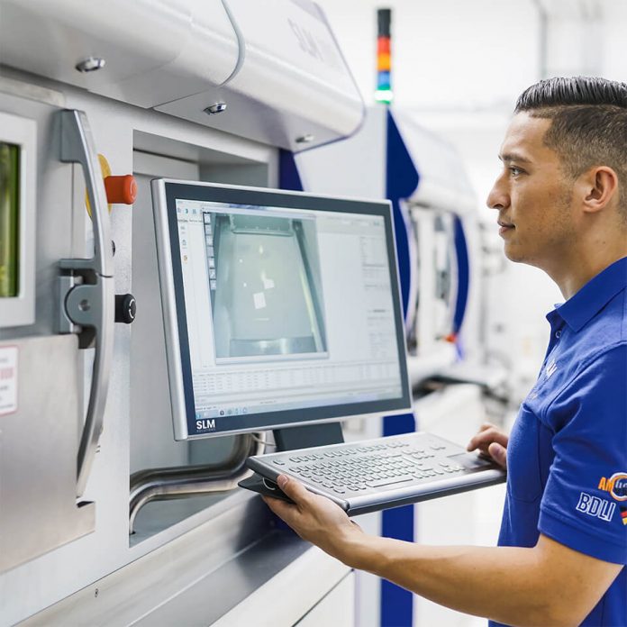 Imagem de um homem operando uma máquina de impressão 3D, com um telão à sua frente.