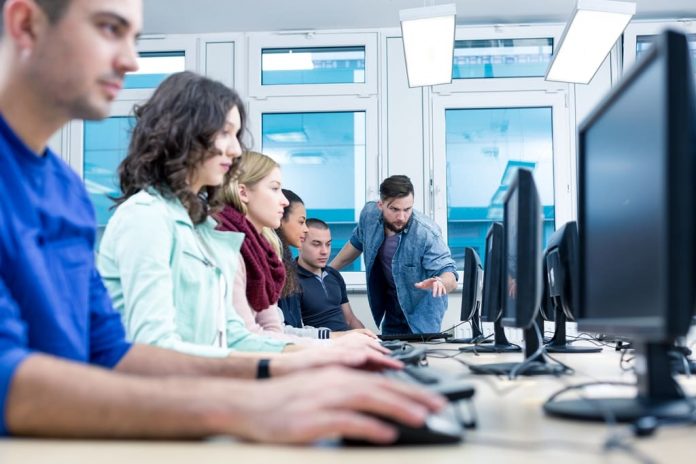 A imagem mostra vários estudantes olhando para a tela de computador. Ao fundo, um professor orienta um dos estudantes.